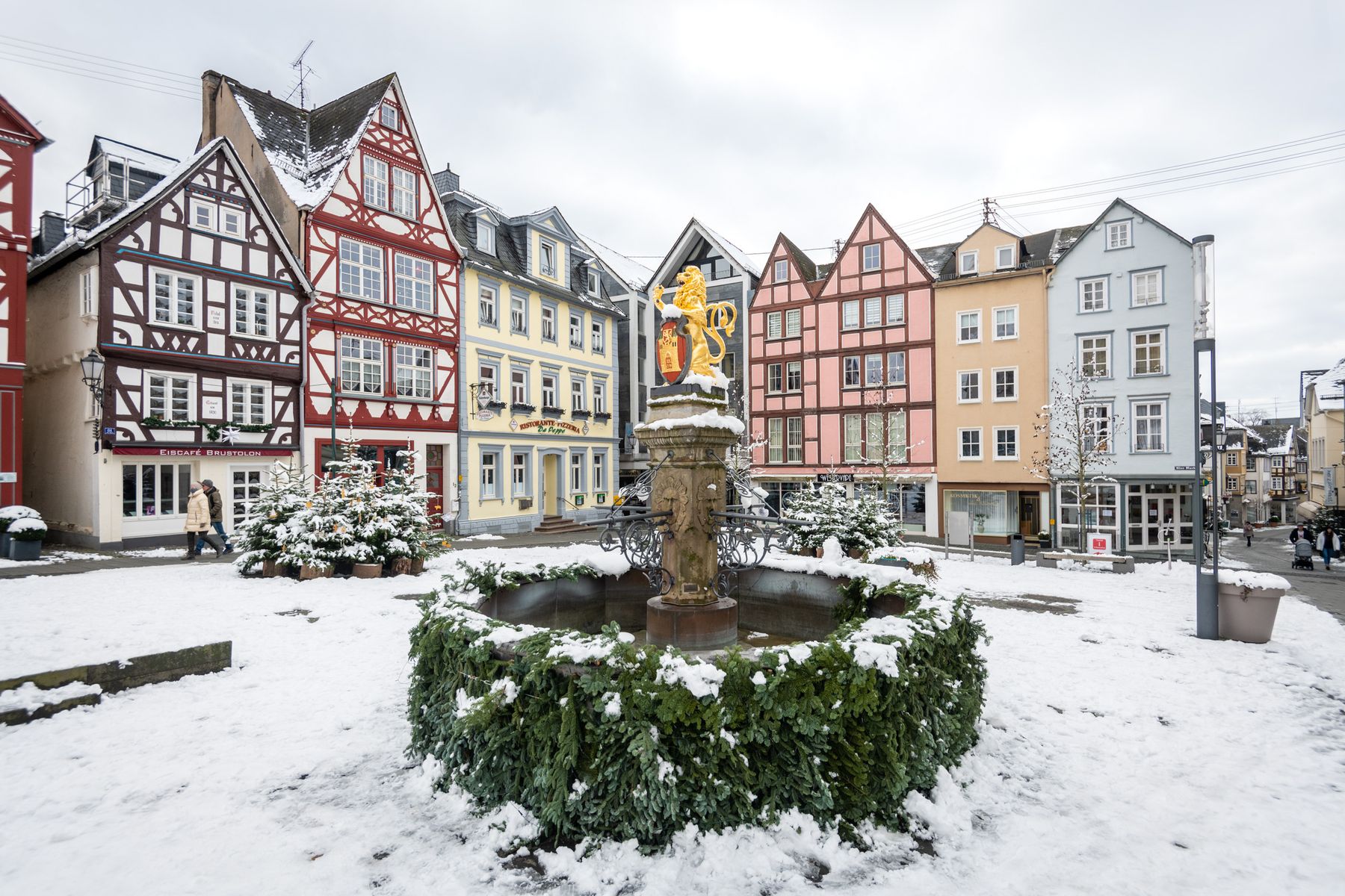 Ein schneebedeckter Stadtplatz mit einem dekorativen Brunnen in der Mitte, umgeben von bunten Fachwerkhäusern. Die Szene hat eine winterliche Atmosphäre mit spazierenden Menschen im Hintergrund.